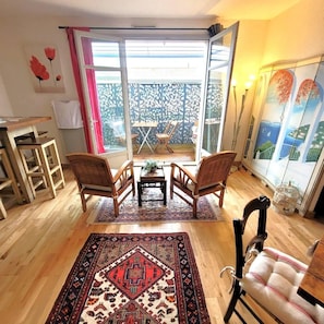 Habitación con piso de madera, alfombra estampada, dos sillas de madera, una mesita y vista a un espacio exterior con cercado decorativo.