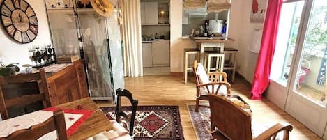 Kodikas olohuone, jossa on puinen ruokapöytä, tuoleja ja kuviollinen matto. Taustalla näkyy keittiö, jossa on ikkuna ja verhot.
