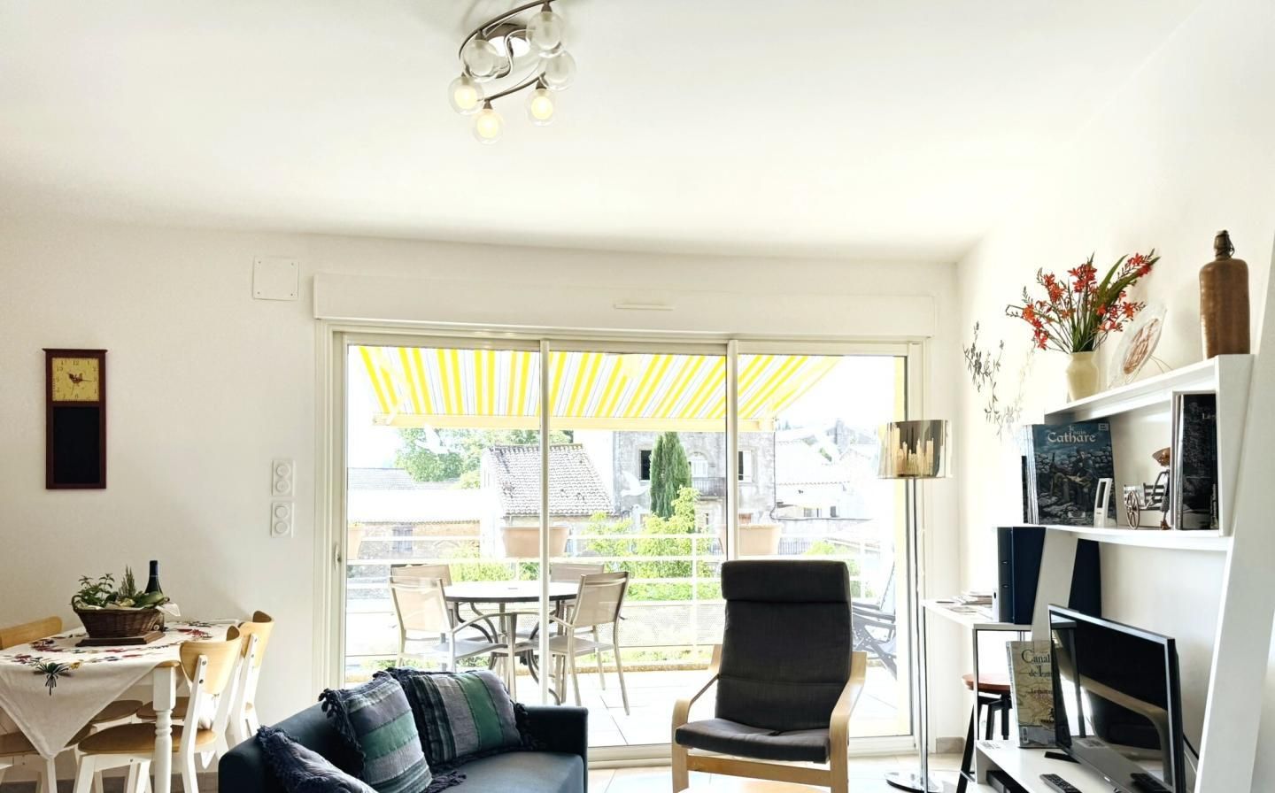 Een woonkamer met een bank, fauteuil, eettafel en stoelen. Er is een tv-meubel met een televisie en een boekenkast. Een gele paraplu staat buiten bij de schuifdeur.