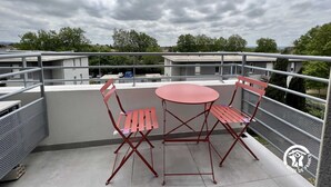 Una terraza en la azotea con una mesa y sillas de metal rojo, una baranda metálica y vistas a edificios y árboles bajo un cielo nublado.
