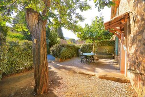 Un camino de grava conduce a una mesa y sillas de madera en un jardín con un gran árbol y un muro de piedra.