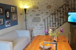 A living room with a sofa, a dining table with a bowl of fruit and flowers, a wall-mounted TV, and a staircase with a stone wall.