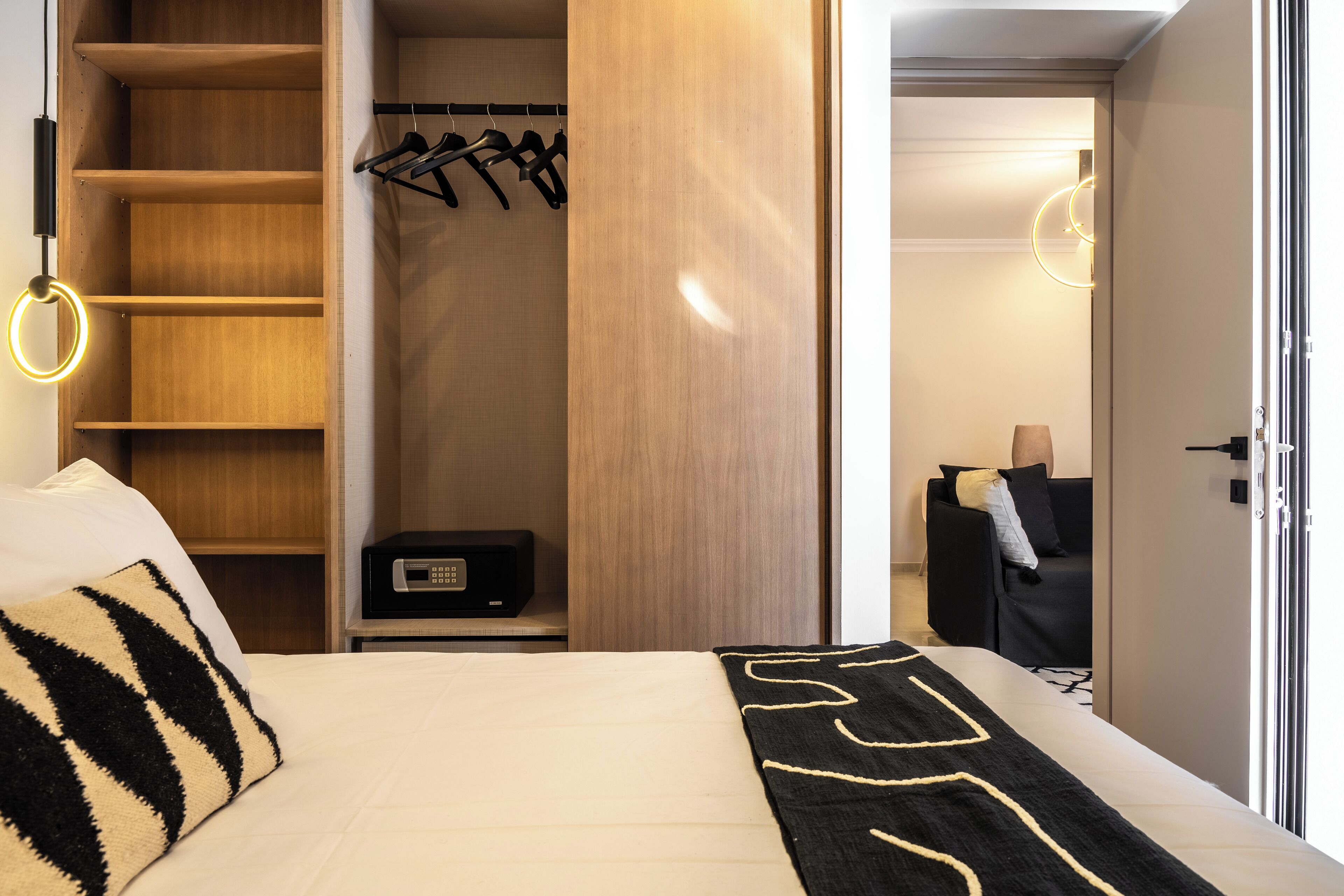 A hotel room with a bed, a wardrobe with hanging clothes, a desk with a radio, and a sofa in the background.