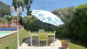 An outdoor patio with a table and chairs, a hanging planter with flowers, and a large umbrella providing shade. - Les Jasmins - PENNAUTIER (PENNAUTIER)