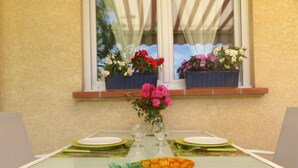 A table set for two with a glass top, two plates, two wine glasses, and a bowl of fruit. There are three flower pots on the windowsill, each with different flowers. - Les Jasmins - PENNAUTIER (PENNAUTIER)