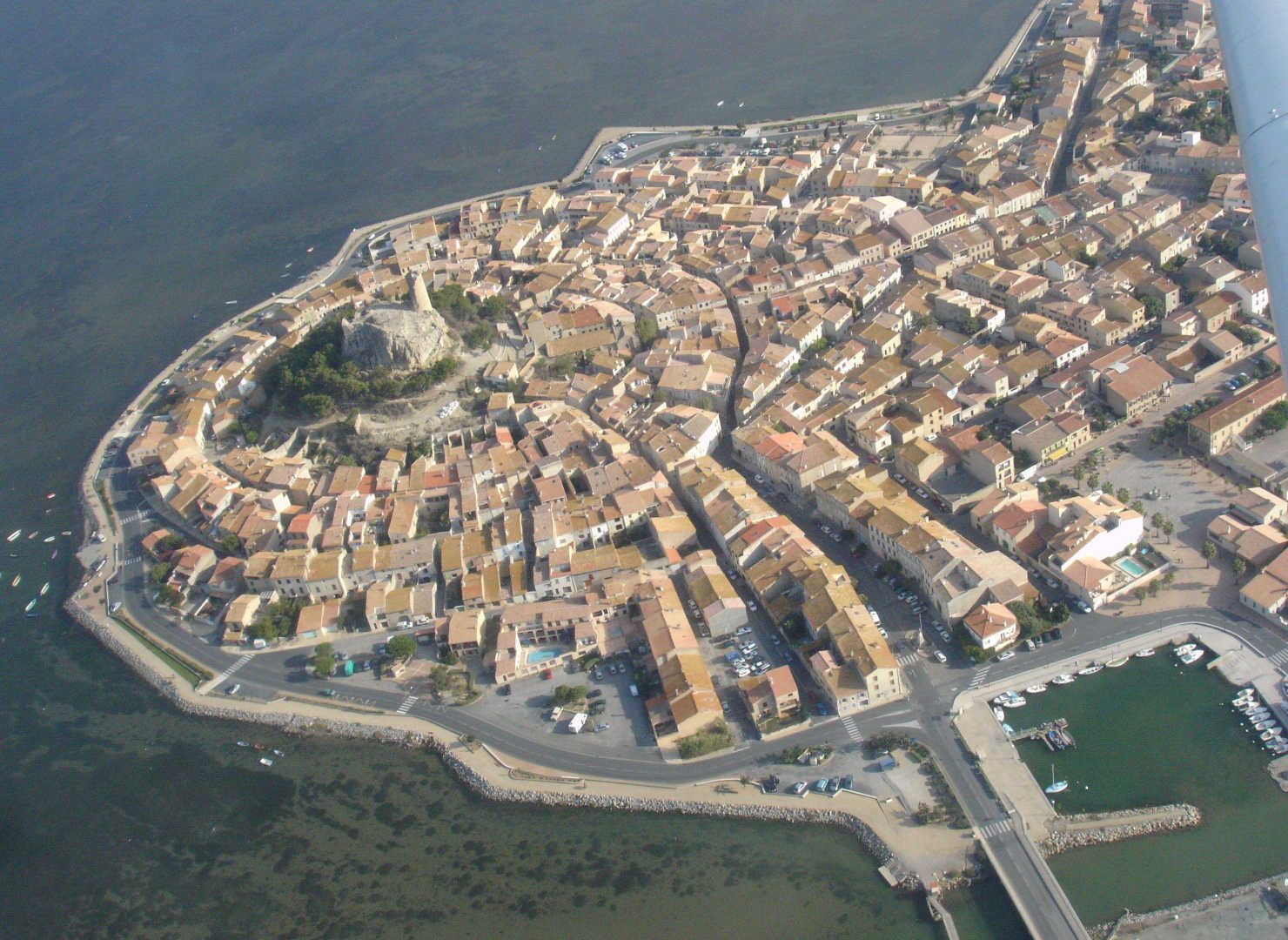 Vue aérienne d’une ville côtière avec une forteresse centrale, des rues étroites et un port de plaisance.