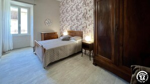 A bedroom with a bed, a wardrobe, a nightstand, a lamp, a window with curtains, and a wall with a floral pattern. - Chez Madou - ROQUEFORT DES CORBIERES (ROQUEFORT DES CORBIERES)