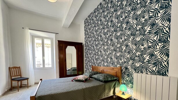 A bedroom with a bed, a chair, a mirror, and a patterned wall. - Chez Madou - ROQUEFORT DES CORBIERES (ROQUEFORT DES CORBIERES)