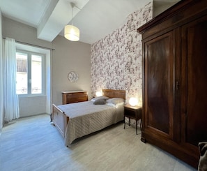 Ein Schlafzimmer mit Bett, Kleiderschrank, Nachttisch, Lampe, einem Fenster mit Vorhängen und einer Wand mit Blumenmuster.