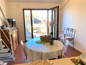 Un coin repas comprenant une table, des chaises, une petite plante et une vue sur une terrasse extérieure, accessible par une porte vitrée.