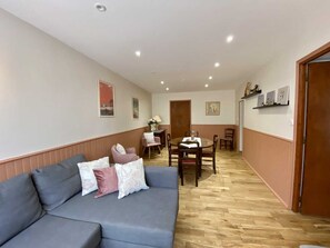 A living room with a sofa, a dining table, and chairs. There are wooden floors and a door leading to another room.