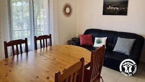 A dining area with a wooden table and chairs, a black sofa with cushions, a decorative wall piece, and a window with sheer curtains.