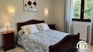 A bedroom with a bed, a nightstand, a lamp, a painting, a window, and a radiator.