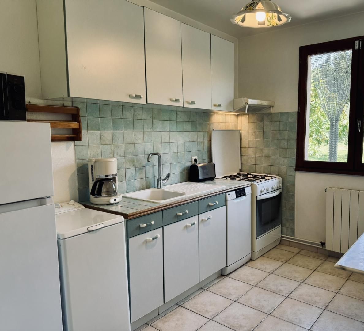 A compact kitchen with white cabinets, a gas stove, a coffee maker, and a window with a view of greenery.