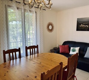 A dining area with a wooden table and chairs, a black sofa with cushions, a decorative wall piece, and a window with sheer curtains.