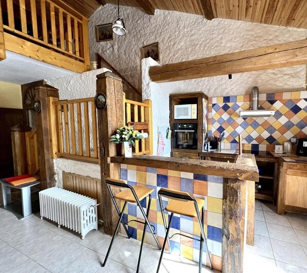 Une cuisine rustique avec un îlot central en bois, des tabourets de bar, un radiateur et un pare-feu en carreaux.