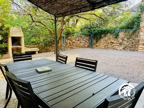 An outdoor patio with a stone wall, a grill, and a table with chairs. - The Courtyard in the Corbières (THEZAN DES CORBIERES)