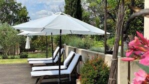 Una terraza con sillones de descanso y una sombrilla grande, rodeada de vegetación y una jardinera de concreto con flores.
