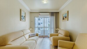 Sala de estar con dos sofás beige, una silla blanca y un ventanal con vistas a edificios.