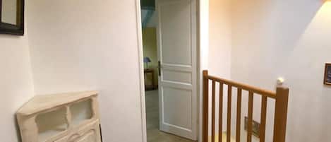Un couloir avec un escalier en bois, une porte blanche donnant sur une autre pièce et un petit meuble.