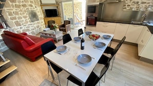 Un coin repas avec une table dressée pour quatre personnes, un canapé rouge, des murs en pierre, une cuisine équipée d’appareils modernes et un téléviseur en arrière-plan.