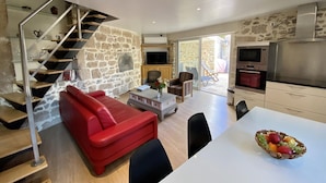Un salon moderne avec un canapé rouge, une table à manger agrémentée d’un panier de fruits, un coin cuisine avec une télévision et un four, et un mur en pierre.