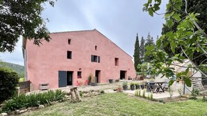 Ein pinkfarbenes Gebäude mit flachem Dach, einem kleinen Garten mit Gras und Pflanzen sowie einer Terrasse mit Gartenmöbeln.