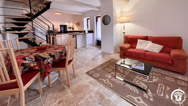 Ein Wohnzimmer mit einem roten Sofa, einem Couchtisch und einem Essbereich mit einem gemusterten Tischläufer. Im Hintergrund ist eine Treppe zu einer Küchenzeile hinaufführend sichtbar.