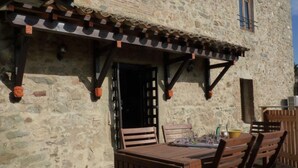 Un bâtiment en pierre avec une table et des chaises en bois à l’extérieur, un toit recouvert de tuiles et une fenêtre avec un cadre en bois.