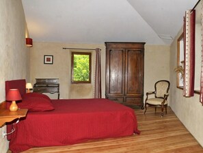 A bedroom with a bed, a wooden wardrobe, a chair, a desk with a lamp, and a window with a view of greenery. - Gîte de la Porte d'Aval (ISSEL)
