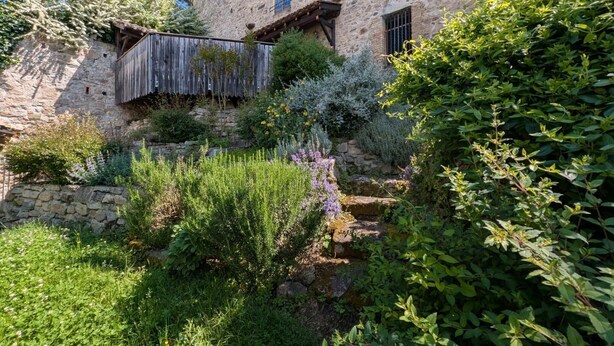 Un bâtiment en pierre, entouré d’une clôture en bois et d’une végétation luxuriante.