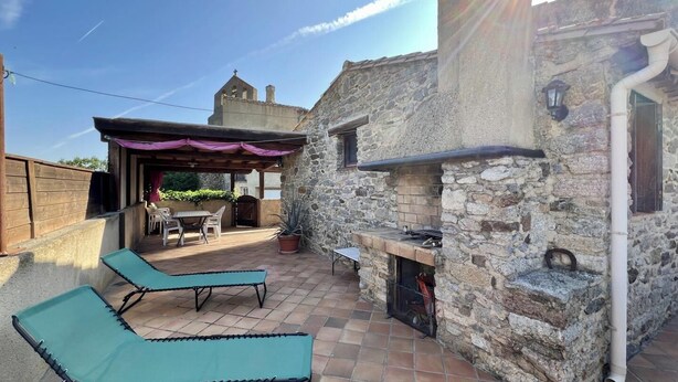 Un bâtiment en pierre doté d’une cheminée, d’un espace extérieur aménagé pour s’asseoir et d’une pergola.