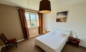 Une chambre à coucher avec un grand lit, une chaise, une table de chevet, une lampe et une fenêtre avec des rideaux.