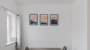 Une pièce comprenant une table à manger et des chaises, une fenêtre avec un rideau, et trois tableaux abstraits encadrés accrochés au mur.
