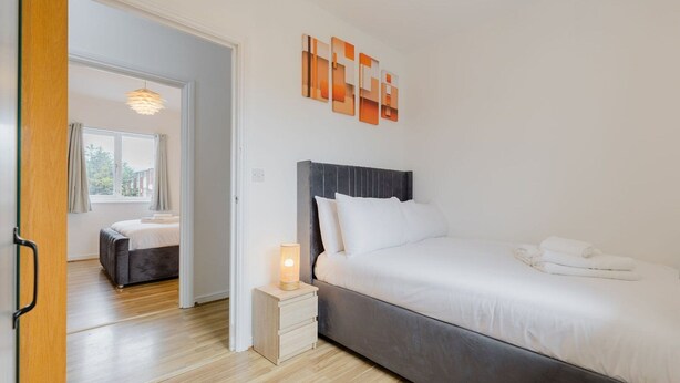 Une chambre moderne avec un grand lit, une table de chevet avec une lampe, et une vue sur une autre pièce par une porte ouverte.
