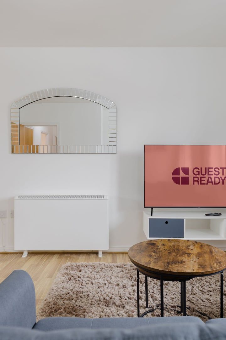 A modern living room with a white console, a round wooden coffee table, a fluffy beige rug, and a flat-screen TV displaying "GUEST READY".