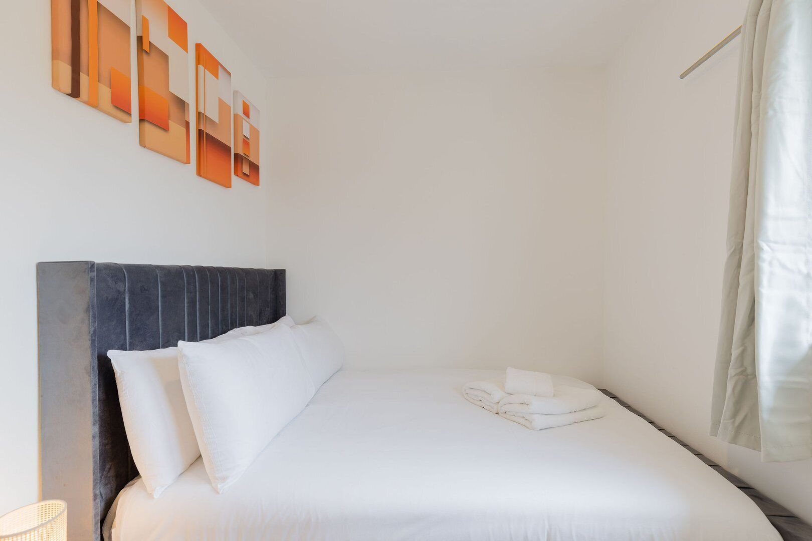 A neatly made bed with white linens and a folded towel, a dark headboard, and abstract wall art.