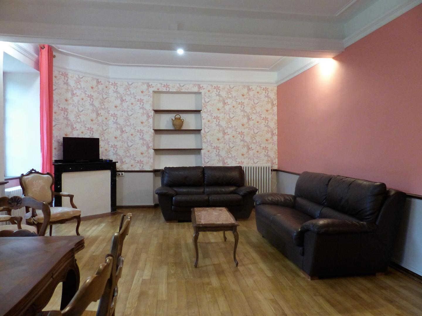 Una sala de estar con piso de madera, un sofá de cuero negro, una mesa de centro de madera, una mesa de comedor de madera con sillas y un radiador.