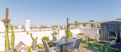 A rooftop patio with a table, chairs, a small house-like structure, and cacti.