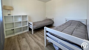 Une chambre avec deux lits simples, une étagère blanche et une lampe.
