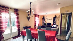 A dining area with a red tablecloth, grey chairs, and a chandelier. There is a TV and a bookshelf in the background.
