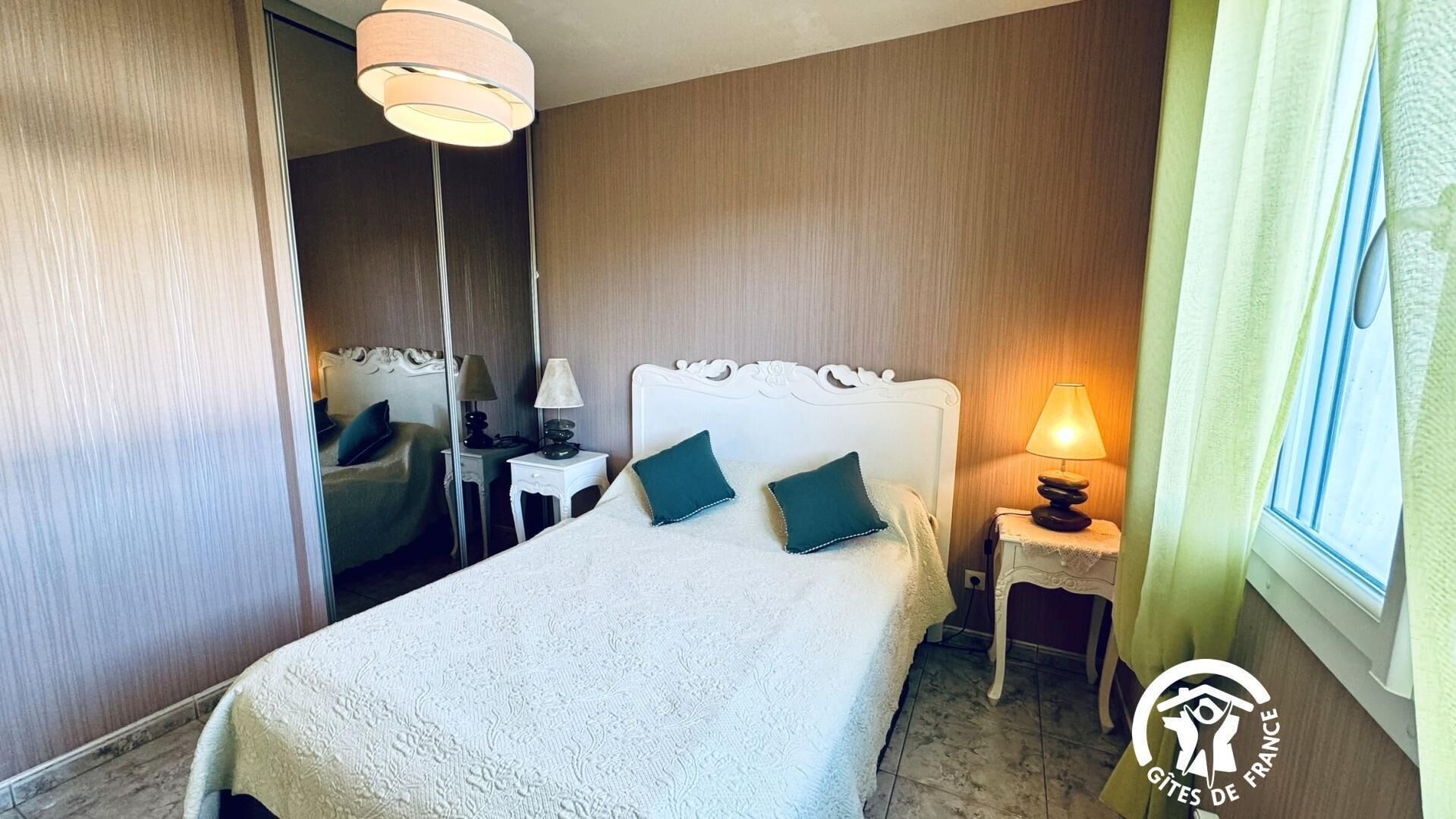 Quarto de hotel com uma cama, duas mesas de cabeceira, uma luminária, um espelho e uma janela com cortinas.