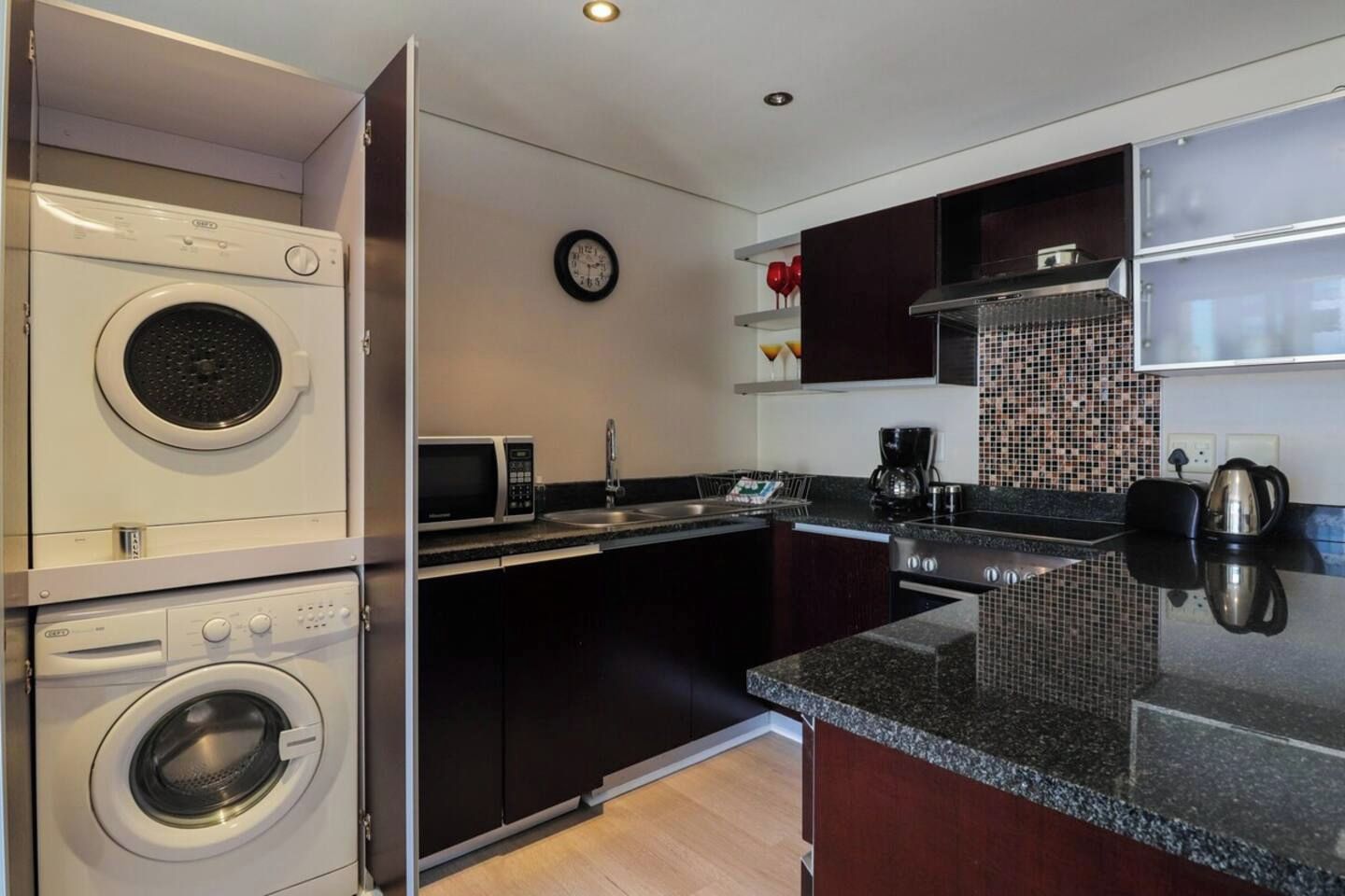 A compact kitchen with a washing machine, microwave, sink, and a clock on the wall.