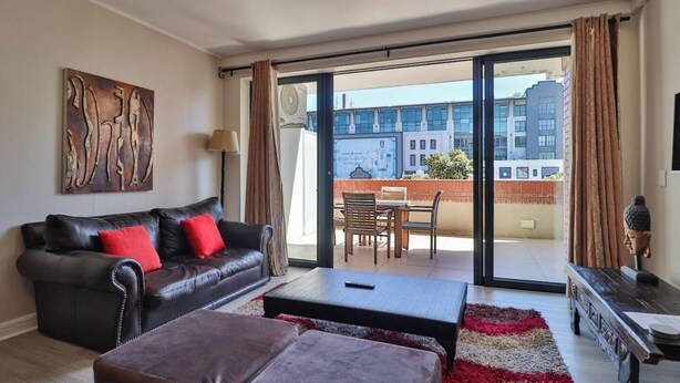 A living room with a leather sofa, a coffee table, and a view of the cityscape through large windows. - De Waterkant 1br|pool, Gym & balcony views by ctha (Cape Town)