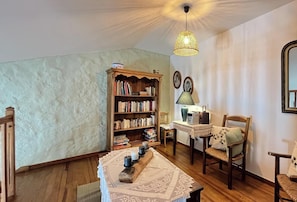 Une pièce chaleureuse avec une bibliothèque en bois, une table recouverte d’une nappe en dentelle, une chaise, une lampe et un luminaire au plafond.