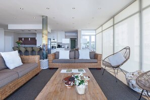 A modern living room with a sofa, armchair, and coffee table, a kitchen in the background, and large windows providing natural light.