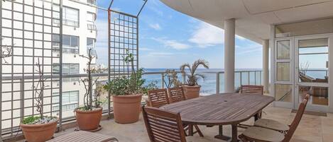 Un patio al aire libre con una mesa y sillas de madera, plantas en macetas y vista al mar.