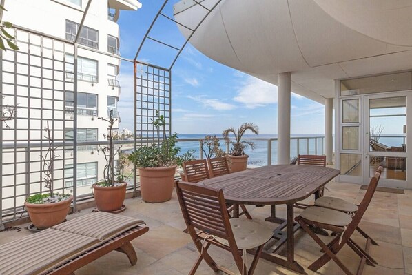 An outdoor patio with a wooden table and chairs, potted plants, and a view of the sea.
