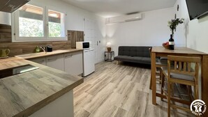 Ett kompakt kök med mikrovågsugn, diskho och träskåp. Det finns en grå soffa och ett matbord med stolar. En platt-TV är monterad på väggen.