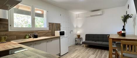 Cocina compacta con microondas, un lavamanos y armarios de madera. Hay un sofá gris y una mesa de comedor con sillas. Un televisor de pantalla plana está montado en la pared.